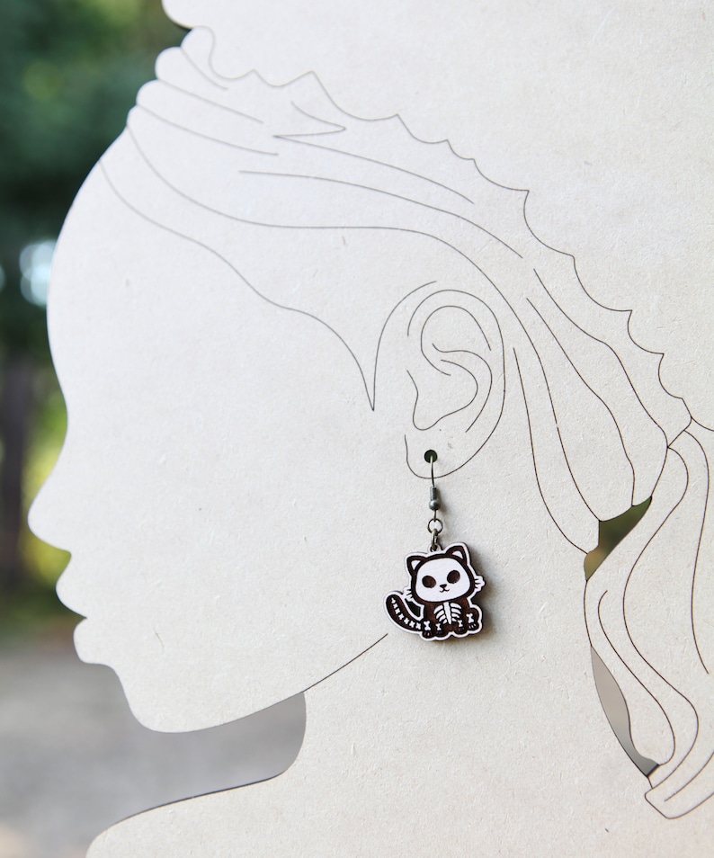 May include: A pair of silver dangle earrings with a black and white cartoon skeleton cat charm.