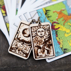 May include: A pair of wooden earrings featuring the tarot cards The Moon and The Sun. The earrings are laser-cut and have a brown wood finish.