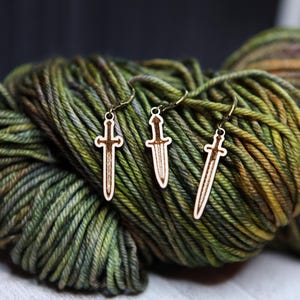 May include: Three wooden sword-shaped stitch markers with gold rings. The markers are on a ball of green yarn.