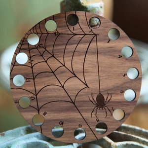 May include: A circular wooden knitting needle gauge with a spider web design and a spider in the center. The gauge has 16 numbered holes for different needle sizes.