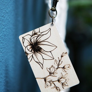 May include: A wooden tag with a black and white floral design, featuring a large magnolia flower and smaller blossoms. The tag is attached to a silver key ring.