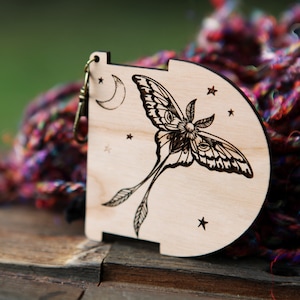 May include: A wooden charm with a laser-etched design of a moth with large eyes, a crescent moon, and stars. The charm is attached to a silver jump ring.