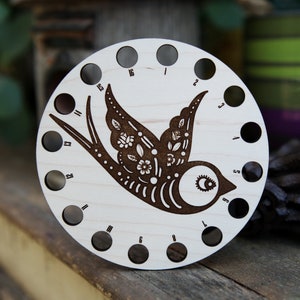May include: A wooden circular yarn bowl with 16 holes around the edge. The bowl is decorated with a laser-cut design of a stylized bird with floral details. The numbers 1 through 16 are printed around the edge of the bowl.