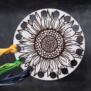 May include: A white wooden sunflower-shaped thread organizer with 16 holes numbered 1 through 16. Three strands of embroidery floss are threaded through the holes.