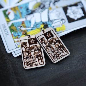 May include: Two wooden earrings with a white background and a brown image of a tarot card, the Death card, with a rider on a horse.