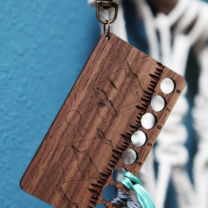 May include: Wooden embroidery floss organizer with a mountain scene design. It has eight holes numbered 4-8 and a metal key ring.  Three strands of floss are threaded through the holes.