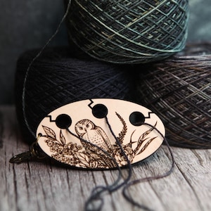 May include: A wooden yarn swift with an engraved owl design, featuring three holes for yarn. The swift is positioned in front of several skeins of yarn in shades of black, gray, and green. The background is a wooden surface.
