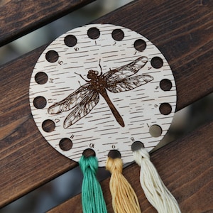 May include: A wooden thread organizer with 15 holes and a dragonfly design. The organizer has four strands of embroidery floss in green, yellow, and white.