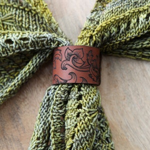 May include: A green and brown knitted scarf with a brown leather ring with a floral design.