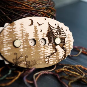 May include: A wooden stitch marker with a whimsical forest scene, featuring a house on stilts, trees, and a moon. The marker has two small, round magnets and the numbers 5 and 6. The edges are scalloped. Yarn is in the background.