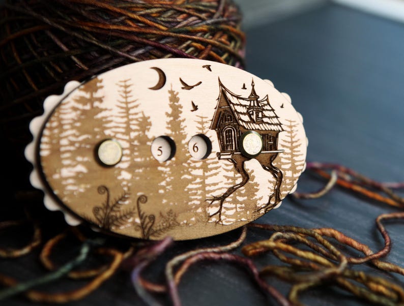 May include: A wooden stitch marker with a whimsical forest scene, featuring a house on stilts, trees, and a moon. The marker has two small, round magnets and the numbers 5 and 6. The edges are scalloped. Yarn is in the background.