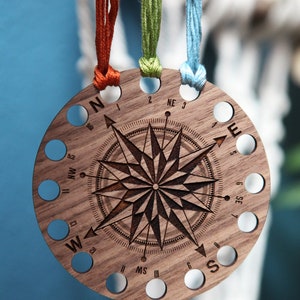 May include: A wooden compass-shaped thread holder with 16 holes for storing embroidery floss. The holder is engraved with a compass rose and the directions North, South, East, and West. Three strands of floss are threaded through the holes.