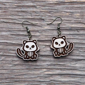 May include: A pair of wooden cat skeleton earrings with brown wood and white paint. The earrings are hanging from silver hooks.