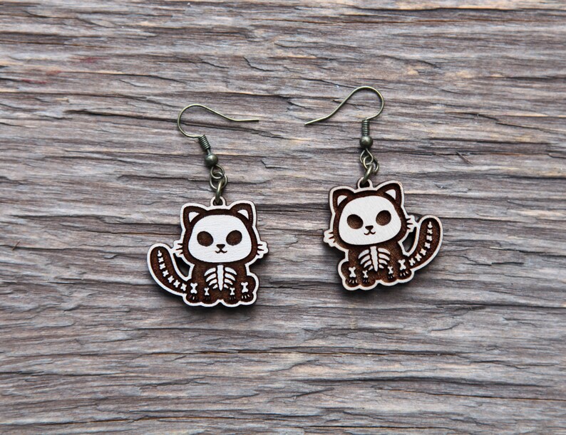 May include: A pair of wooden cat skeleton earrings with brown wood and white paint. The earrings are hanging from silver hooks.