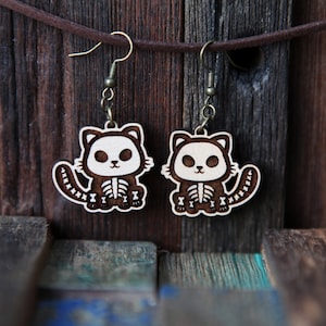 May include: A pair of wooden cat skeleton earrings with a brown finish and white details. The earrings are hanging from a brown leather cord against a wooden background.