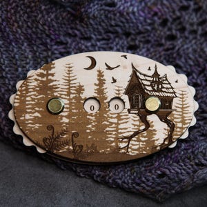 May include: A wooden oval-shaped knitting row counter with a forest and house design. The counter has two brass-colored magnets and the numbers "0 0" etched into the wood. The background features a forest scene with a crescent moon and birds.