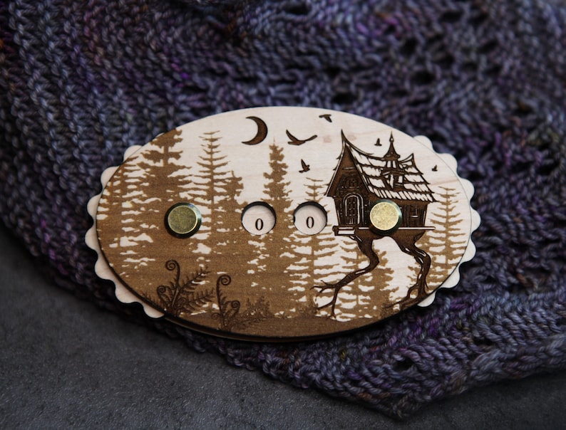 May include: A wooden oval-shaped knitting row counter with a forest and house design. The counter has two brass-colored magnets and the numbers "0 0" etched into the wood. The background features a forest scene with a crescent moon and birds.