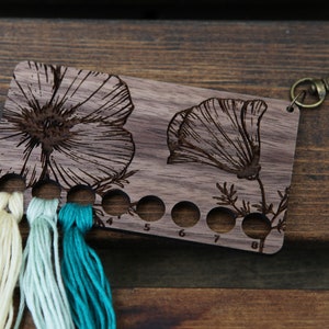 May include: Wooden thread organizer with a floral design and eight numbered holes for storing embroidery floss. The organizer has a metal clasp for attaching to a bag or keychain. Three strands of floss are threaded through the holes.
