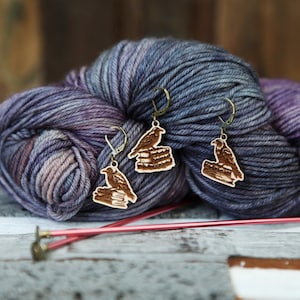 May include: A pair of wooden earrings with a raven perched on a stack of books. The earrings are hanging from a skein of purple and blue yarn.