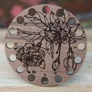 May include: A wooden circular stitch holder with 16 holes numbered 1 through 16. The holder is decorated with an engraved floral design of fuchsia flowers and leaves.