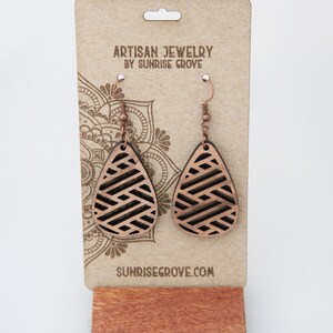 May include: A pair of wooden teardrop earrings with a geometric pattern. The earrings are on a brown card with the text "Artisan Jewelry by Sunrise Grove" and "Sunrisegrove.com".