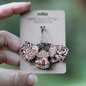 May include: A set of six wooden mushroom stitch markers with a metal ring. The markers are decorated with different mushroom designs and the text "Shroom Queens" and "Stitch Markers".