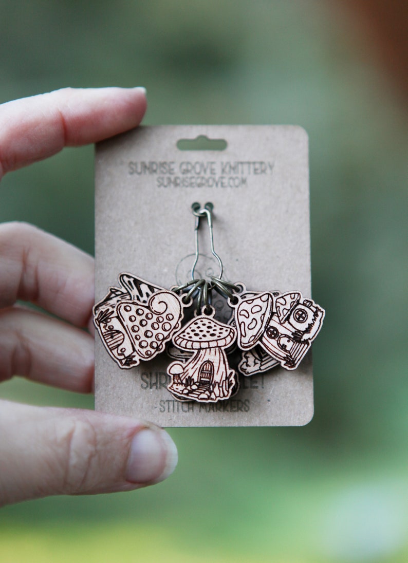 May include: A set of six wooden mushroom stitch markers with a metal ring. The markers are decorated with different mushroom designs and the text "Shroom Queens" and "Stitch Markers".