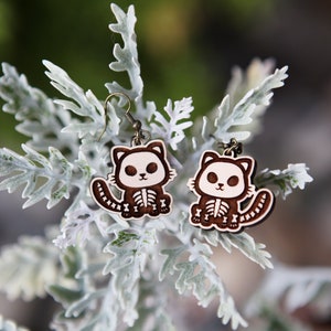May include: A pair of wooden earrings featuring a cute cartoon cat with a skeleton design. The cat is brown with white accents and a white skeleton. The earrings are hanging from a silver hook.