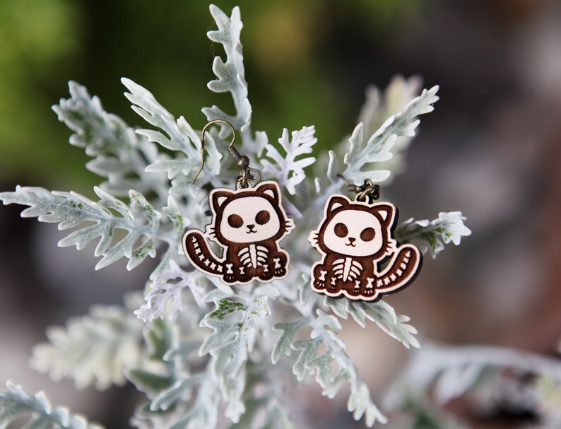 May include: A pair of wooden earrings featuring a cute cartoon cat with a skeleton design. The cat is brown with white accents and a white skeleton. The earrings are hanging from a silver hook.