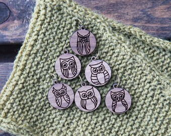 May include: Six wooden owl stitch markers with a brown wood finish. The owls are all different designs and have black eyes. The markers are on a green knitted fabric.