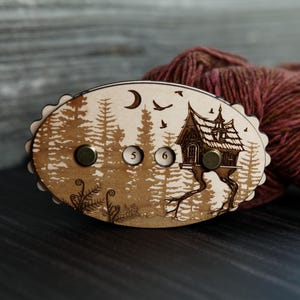 May include: Wooden yarn swift with a forest and house design. The oval-shaped swift features a detailed engraving of a forest scene with a house, moon, and birds. It has two brass knobs and the numbers 5 and 6.