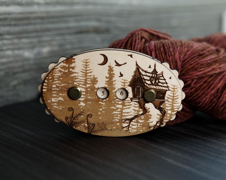 May include: Wooden yarn swift with a forest and house design. The oval-shaped swift features a detailed engraving of a forest scene with a house, moon, and birds. It has two brass knobs and the numbers 5 and 6.