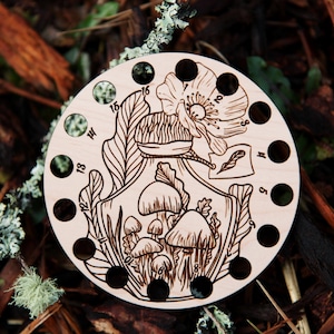 May include: Wooden circular stitch marker with 18 holes and a design of a poppy flower, mushrooms, and leaves. The numbers 8 through 18 are printed around the edge.