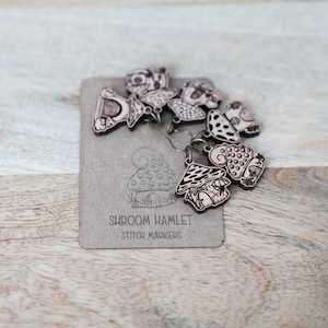 May include: A set of wooden mushroom stitch markers with a brown and white color scheme. The markers are shaped like mushrooms with different designs and are labeled "Shroom Hamlet - Stitch Markers".