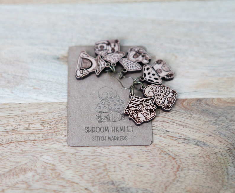 May include: A set of wooden mushroom stitch markers with a brown and white color scheme. The markers are shaped like mushrooms with different designs and are labeled "Shroom Hamlet - Stitch Markers".