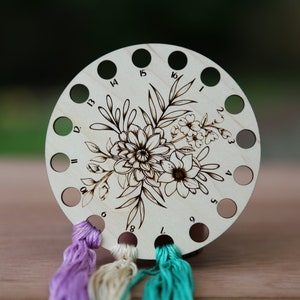 May include: Wooden thread organizer with 16 holes and a floral design. The organizer has numbers 1 through 16 around the edge. Three strands of embroidery floss are threaded through the holes.