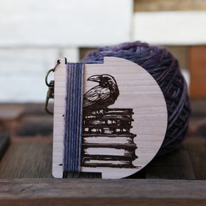 May include: Wooden yarn bowl with a raven perched on a stack of books. The bowl is decorated with a laser-engraved design and has a blue yarn wrapped around it.