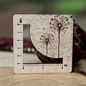 May include: Wooden ruler with a dandelion design and a measurement scale in inches and centimeters. The ruler is 5 inches long and has a hole for a keychain.