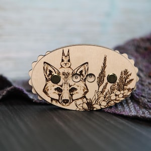 May include: Wooden fox and rabbit design stitch marker with two gold metal beads. The marker is oval shaped and has a wood grain finish.