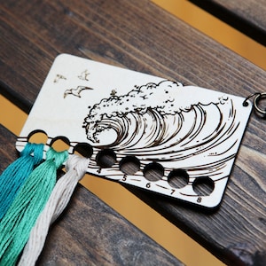 May include: Wooden thread organizer with a wave design and seagulls. It has eight numbered holes for holding embroidery floss. The organizer has a metal key ring attached.