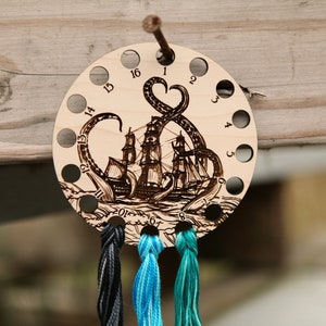May include: Wooden thread holder with 16 holes, featuring a laser-etched design of an octopus attacking a ship. The holder has three strands of yarn in black, teal, and turquoise colors.