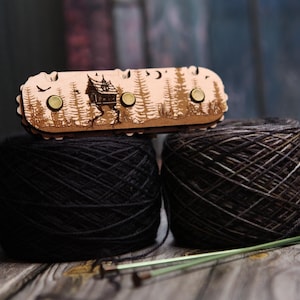 May include: A wooden yarn needle case with a laser-cut forest and house design, resting on two balls of dark yarn. The case has three gold-colored closures. Knitting needles are visible in the foreground.