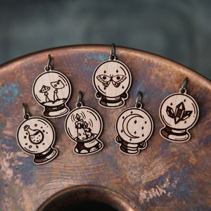 May include: Six wooden charms shaped like a crystal ball with different etched designs inside. The designs include a mushroom, a butterfly, a candle, a crescent moon, a crystal, and a potion bottle.