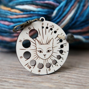 May include: A wooden yarn bowl with a cat face design and a needle holder. The bowl has 10 holes for yarn and is numbered 0 to 6. The bowl is attached to a metal ring.