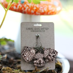 May include: A set of five wooden mushroom stitch markers with a house design. The markers are attached to a silver safety pin and are labeled "Sunrise Grove Knittery - Stitch Markers".