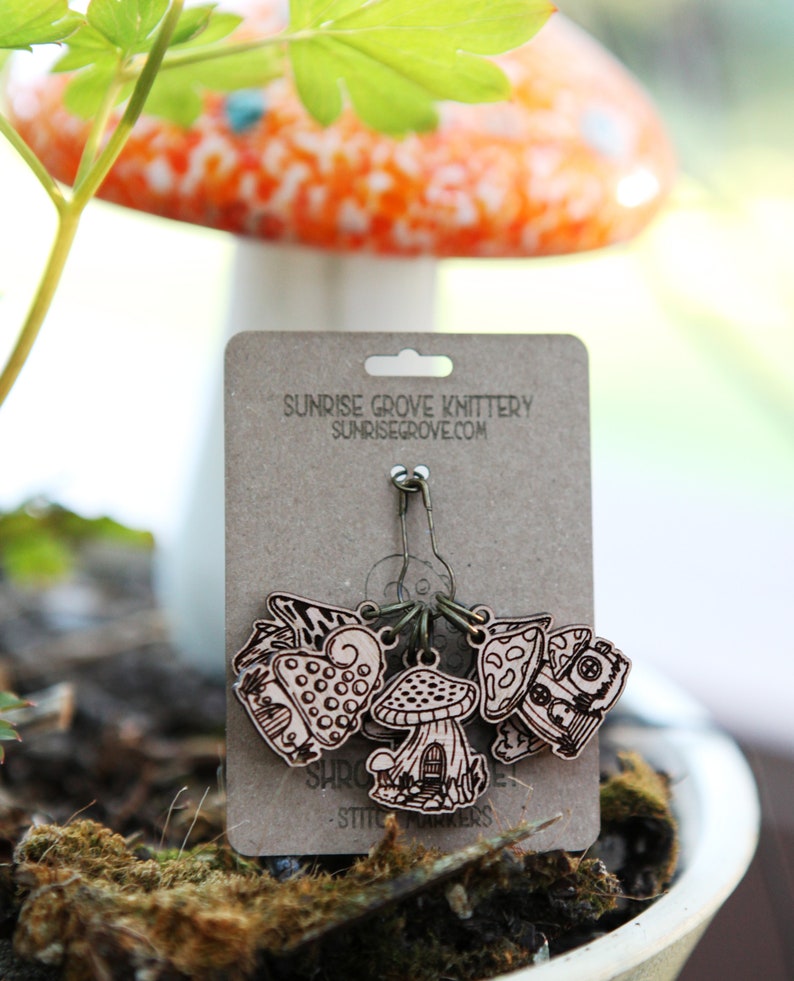May include: A set of five wooden mushroom stitch markers with a house design. The markers are attached to a silver safety pin and are labeled "Sunrise Grove Knittery - Stitch Markers".