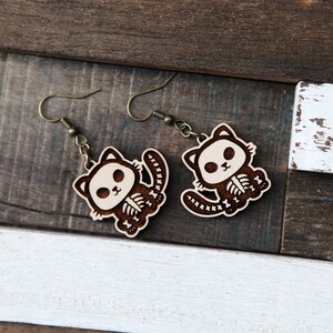 May include: A pair of wooden earrings featuring a cute cat skeleton design. The earrings are brown with white details and have a dangling hook style.
