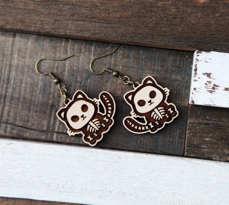 May include: A pair of wooden earrings featuring a cute cat skeleton design. The earrings are brown with white details and have a dangling hook style.