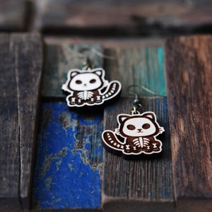 May include: A pair of wooden earrings featuring a skeleton cat design. The earrings are brown with white details and have a silver hook.