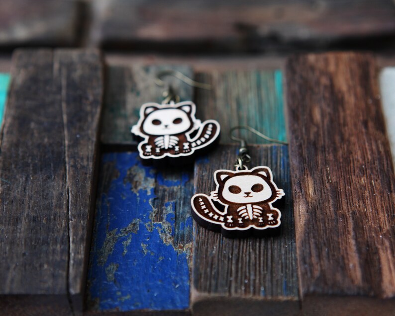 May include: A pair of wooden earrings featuring a skeleton cat design. The earrings are brown with white details and have a silver hook.